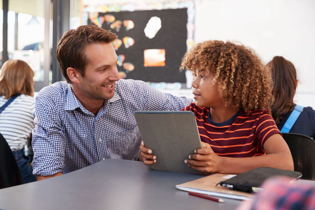 male-teacher-helping-student-at-their-desk-in-a-classroom male teacher helping student at their desk in a classroom