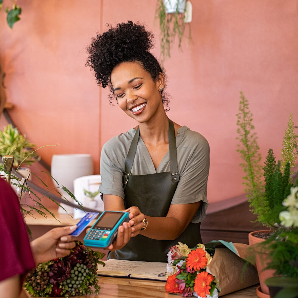 Smiling florist holding card reader machine at counter with customer paying with credit card. Young african american florist shop assistant holding payment machine while buyer purchase a bunch flower.